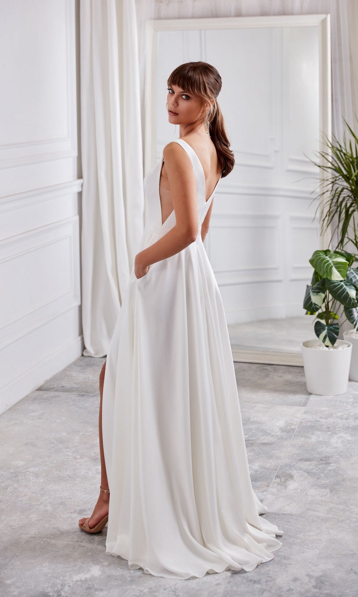 A woman in the Long White Dress: Aerie Couture 2007, a sleeveless A-line formal dress with pockets, stands indoors looking over her shoulder. A potted plant and a mirror are visible in the background.